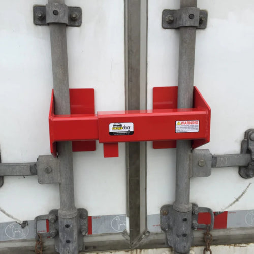 A red HD Cargo Door Lock from Container Nut clamped onto the metal handles of a closed door. The device features warning labels and secures the door by preventing the handles from being moved. The background shows the white surface of the vehicle.