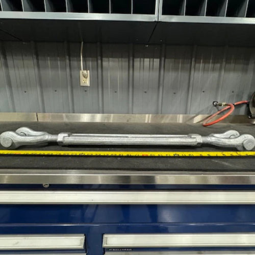 A Container Nut Galvanized Turnbuckle with hooks on both ends is placed on a workbench in front of a wall with metal panels. A yellow measuring tape lies parallel to the turnbuckle. The background shows organized shelves and an electrical outlet with cables.