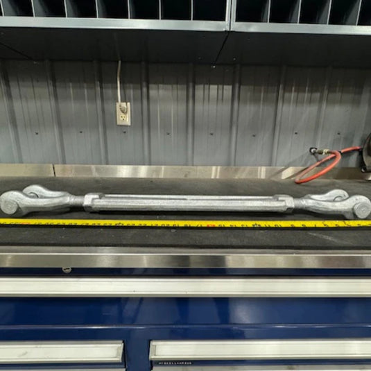 A Container Nut Galvanized Turnbuckle with hooks on both ends is placed on a workbench in front of a wall with metal panels. A yellow measuring tape lies parallel to the turnbuckle. The background shows organized shelves and an electrical outlet with cables.