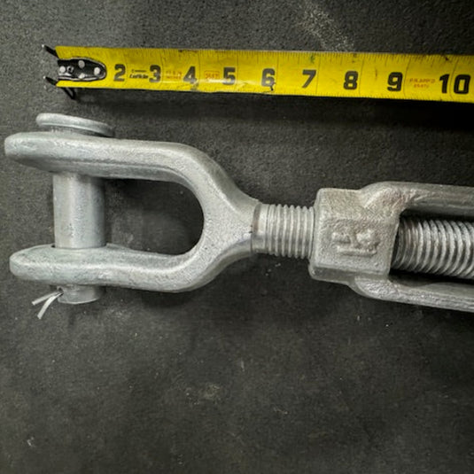 A close-up image of the Galvanized Turnbuckle by Container Nut next to a yellow measuring tape displaying measurements in inches from 2 to 11. The turnbuckle, made of metal, features a threaded rod and a jaw end fitting. The background is a dark, textured surface.