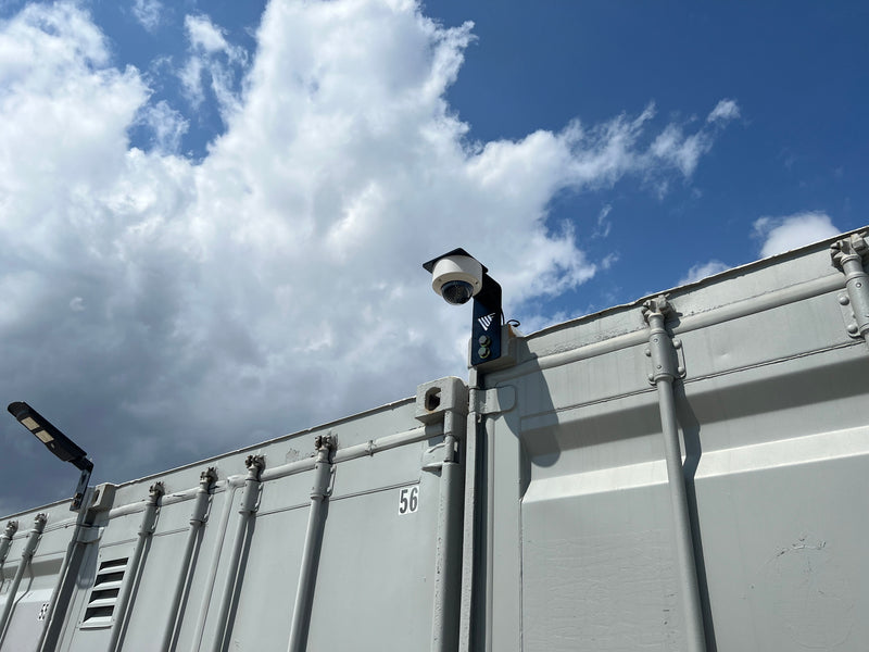 Load image into Gallery viewer, Under a partly cloudy sky, a security camera is mounted on the side of a large gray metal container. It&#39;s mounted by nuts from Container Nut. A streetlight can be seen at the left edge of the image.
