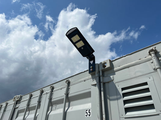 Under a partly cloudy sky, a security camera is mounted on the side of a large gray metal container. It's mounted by nuts from Container Nut.